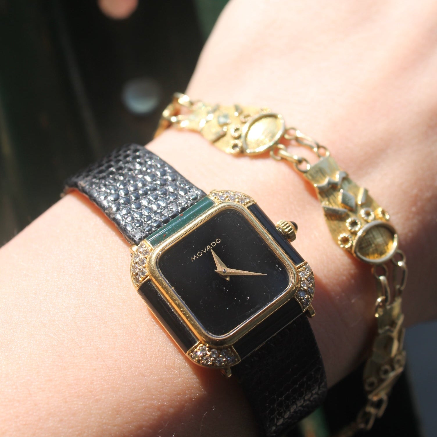 Hand wearing a black square watch with gold accents and multiple bracelets.