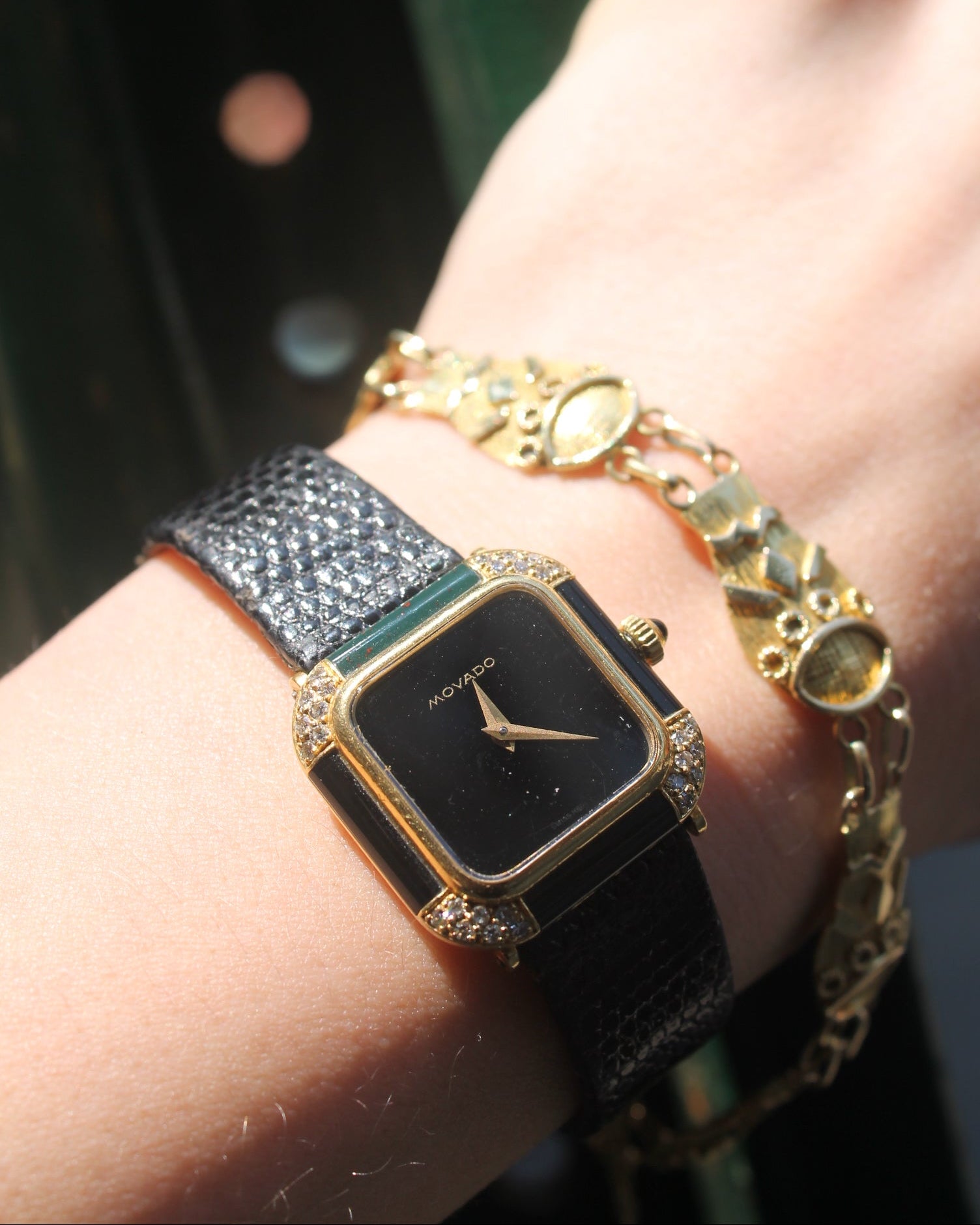 Hand wearing a black square watch with gold accents and multiple bracelets.